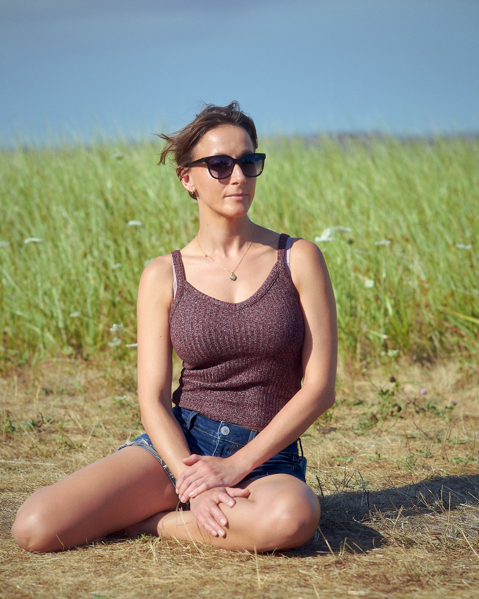 Woman sitting in the grass in the sunshine in a tank top and shorts with heart shaped necklace
