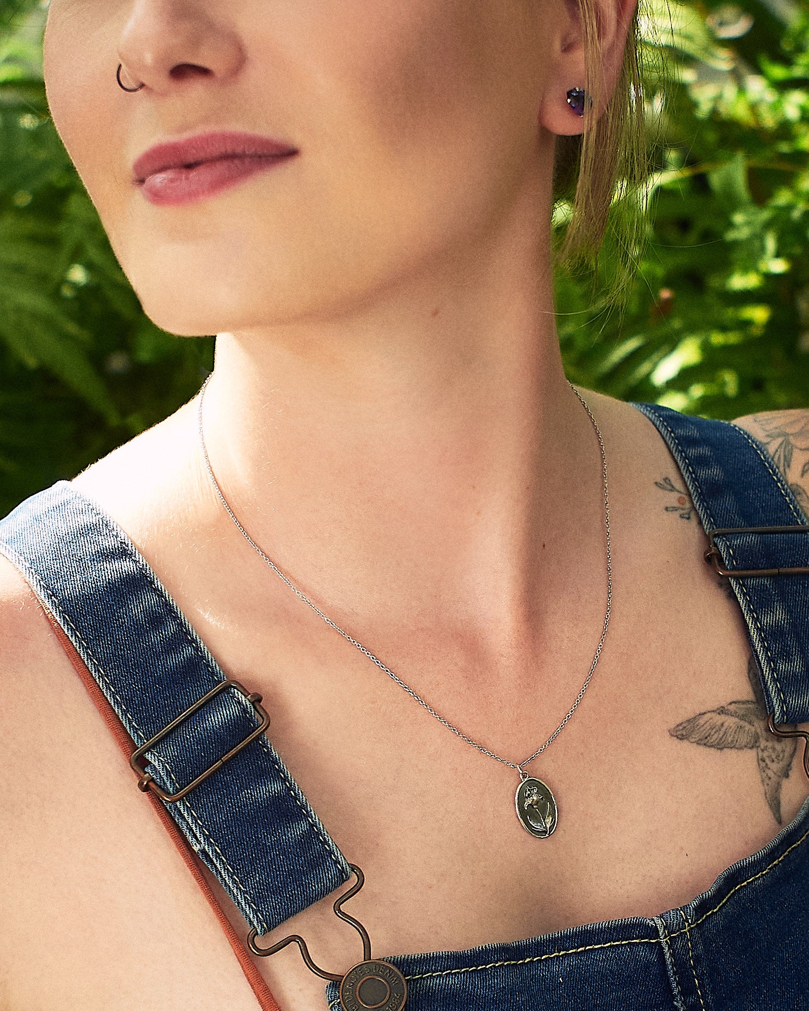 A close-up lifestyle shot of a woman wearing a delicate silver chain necklace with an oval pendant featuring a cameo-style floral, styled with blue denim overalls against a blurred green foliage background.