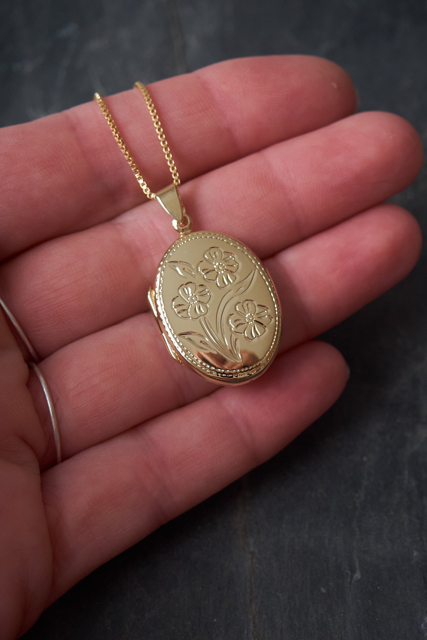 Gold locket with floral design held in a hand against a dark background