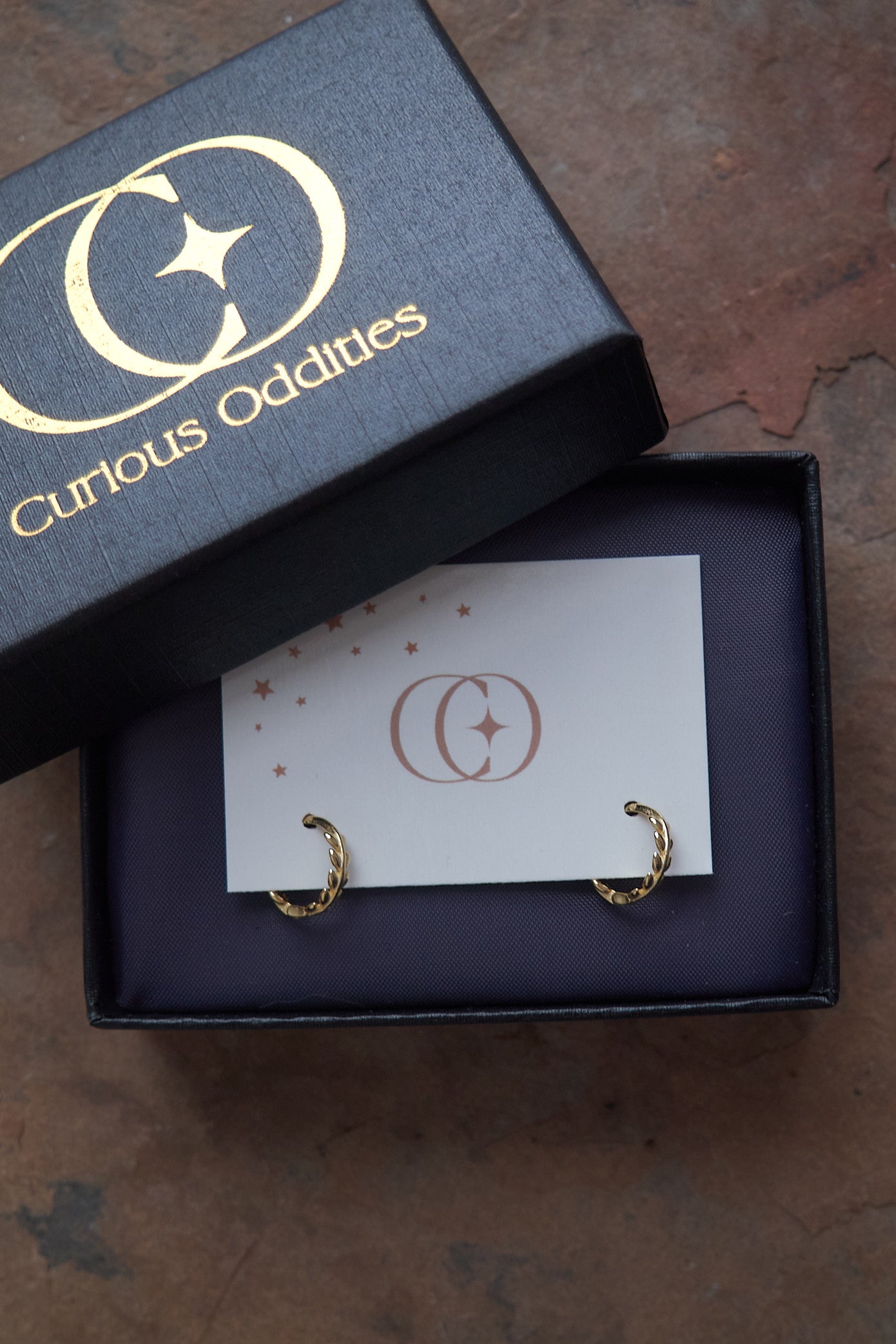 Gold hoop earrings in a 'Curious Oddities' box on a stone surface