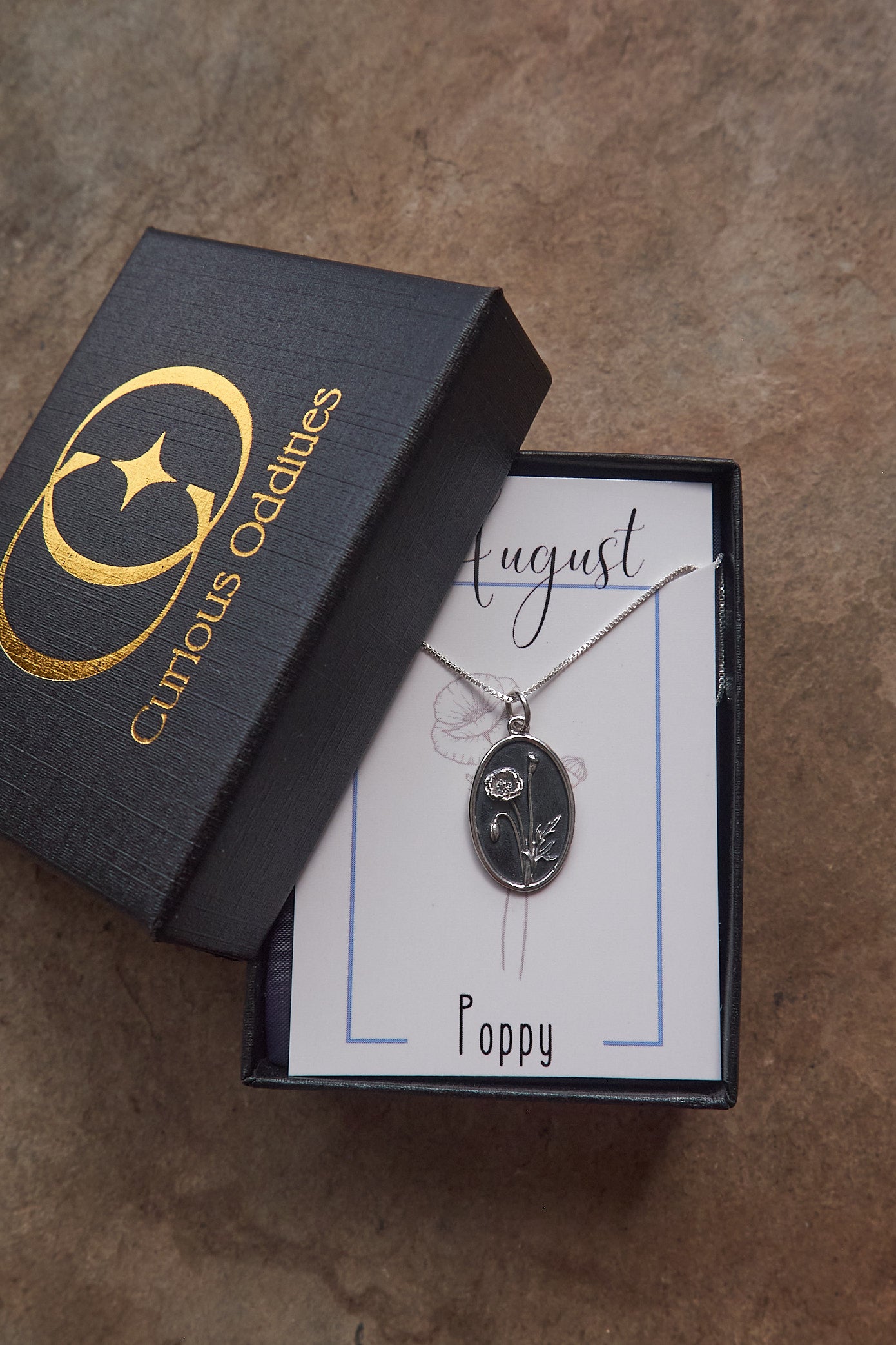 Necklace with a floral pendant on a card inside a 'Curious Oddities' box on a brown slate surface