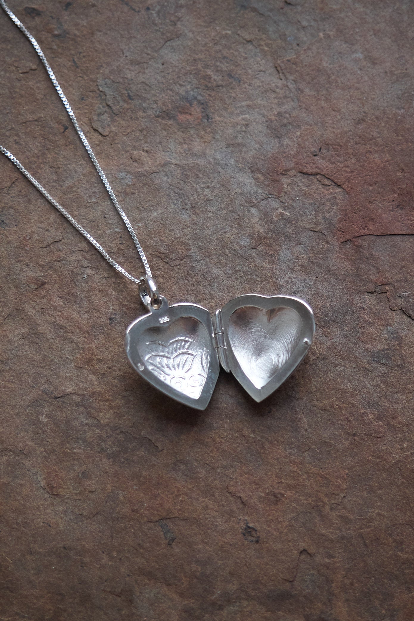 Silver heart-shaped locket on a chain shown open on a brown stone background