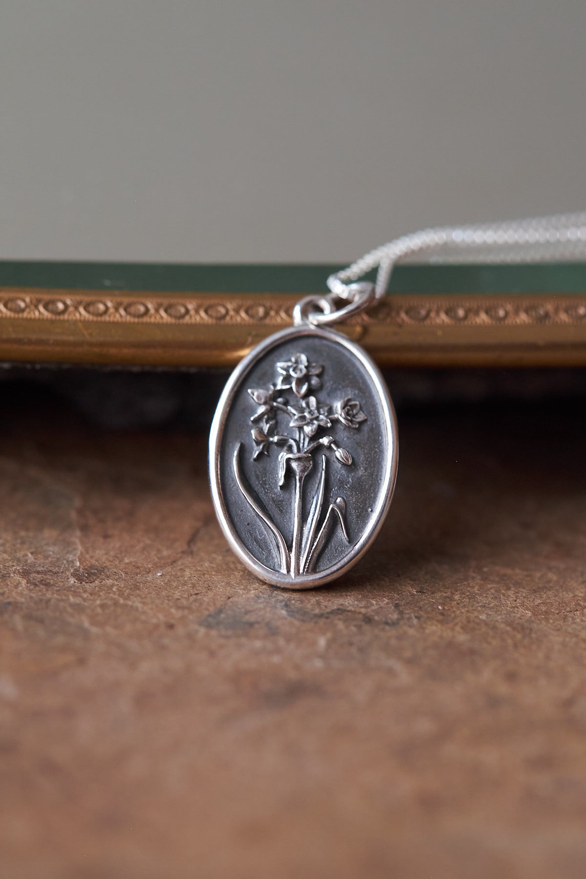 An oval silver pendant necklace featuring an embossed daffodil design, resting on a textured brown surface with a blurred decorative frame in the background.