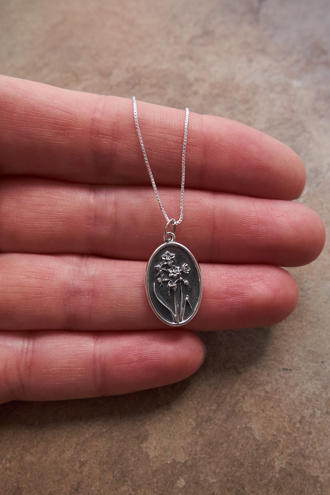 Silver necklace with a pendant featuring a floral design held between fingers on a brown surface.