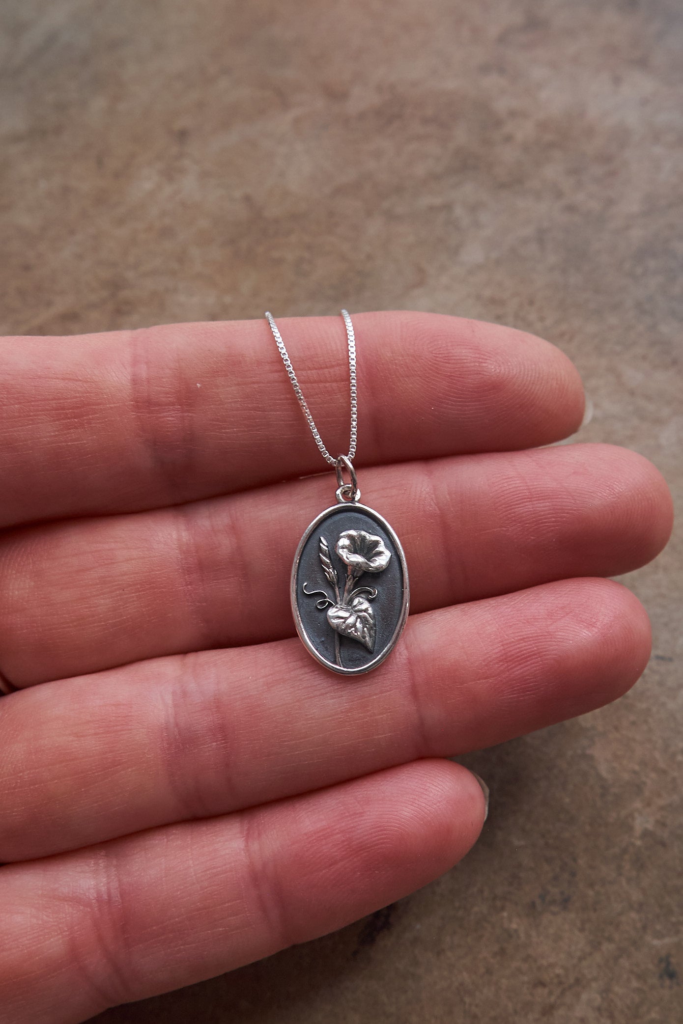 Silver necklace with an oval pendant featuring a floral design held in a hand on a brown slate surface.