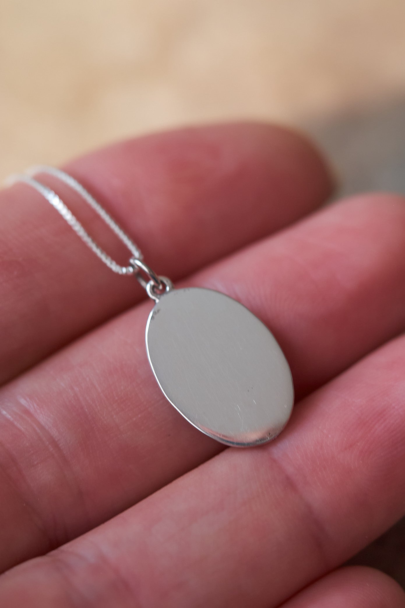 Silver necklace with the smooth back of an oval pendant held in hand on a blurred background