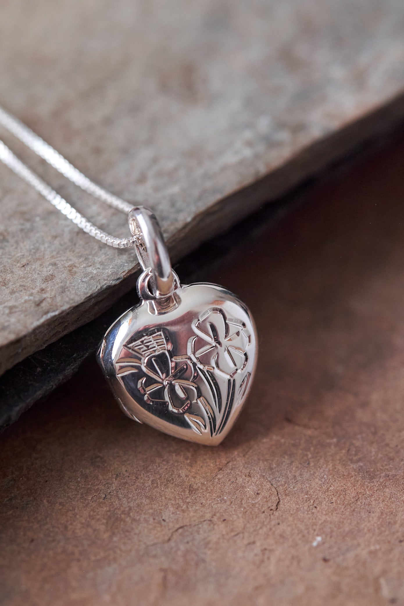 Silver heart-shaped locket pendant with floral design on a stone surface