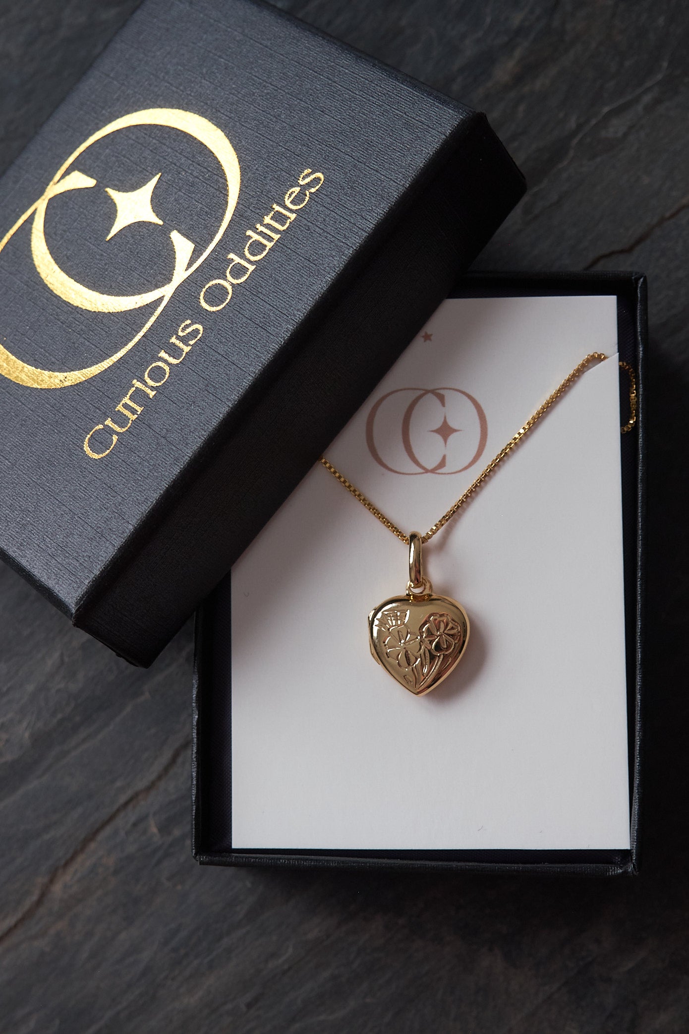 Gold heart-shaped locket pendant necklace in a 'Curious Oddities' box on a dark slate surface