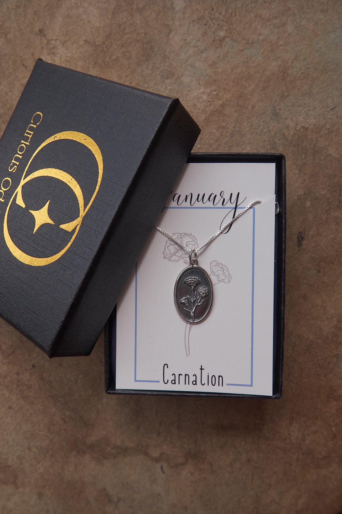 Silver carnation birth flower necklace on a card, next to an open box with 'Curious Oddities' branding.
