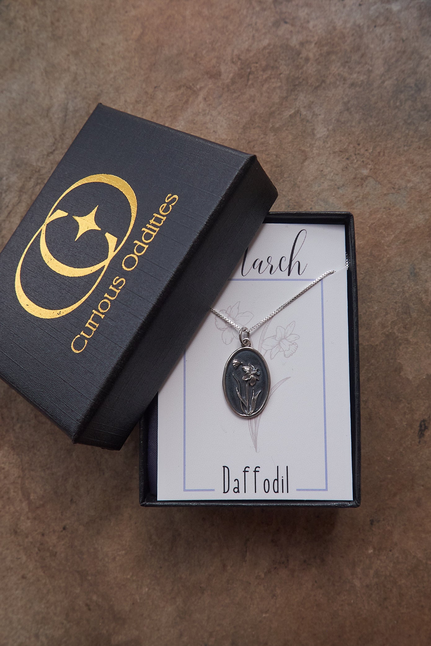 Silver necklace with floral pendant in a 'Curious Oddities' box on a brown slate surface