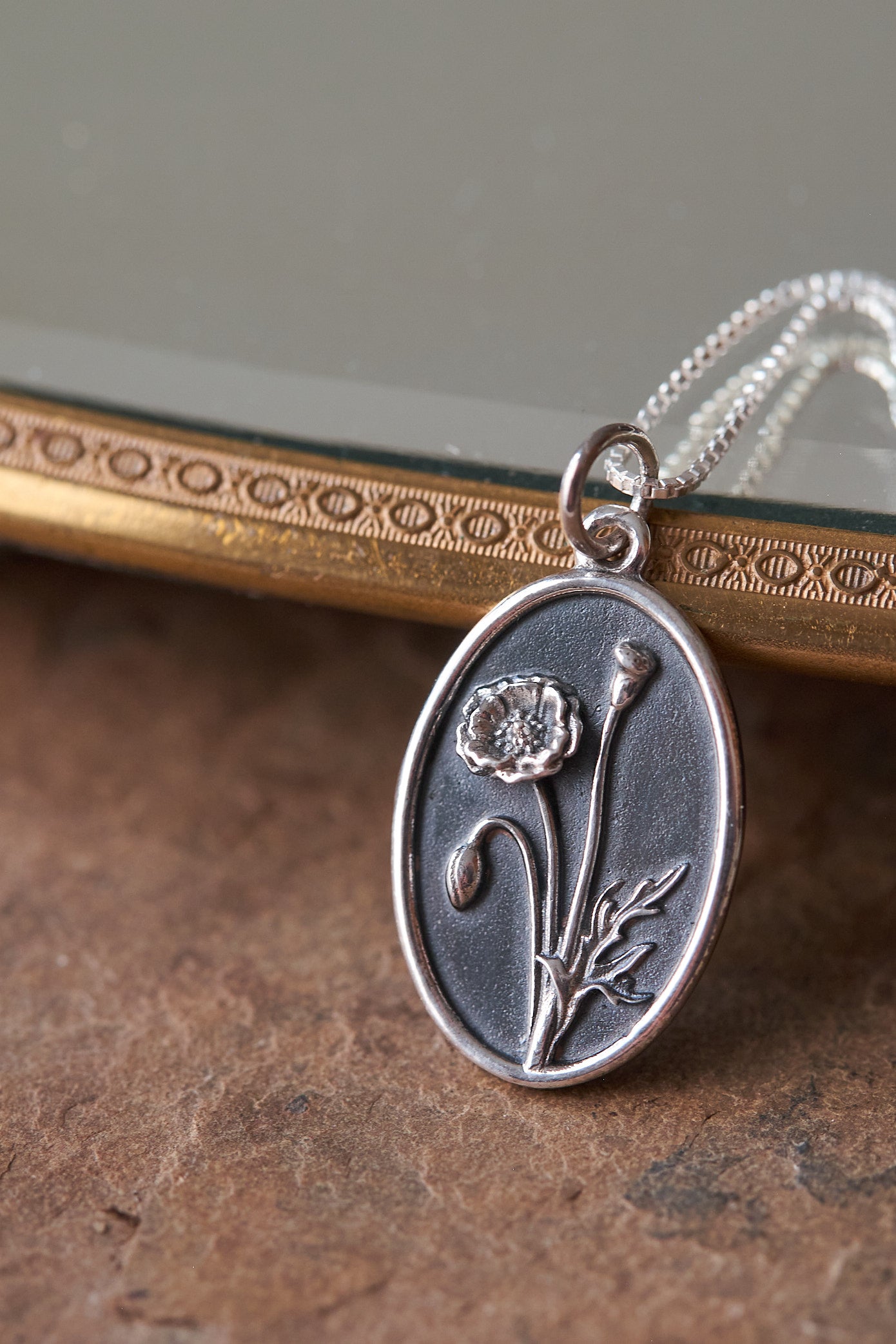 Silver poppy pendant on a chain against a reflective surface