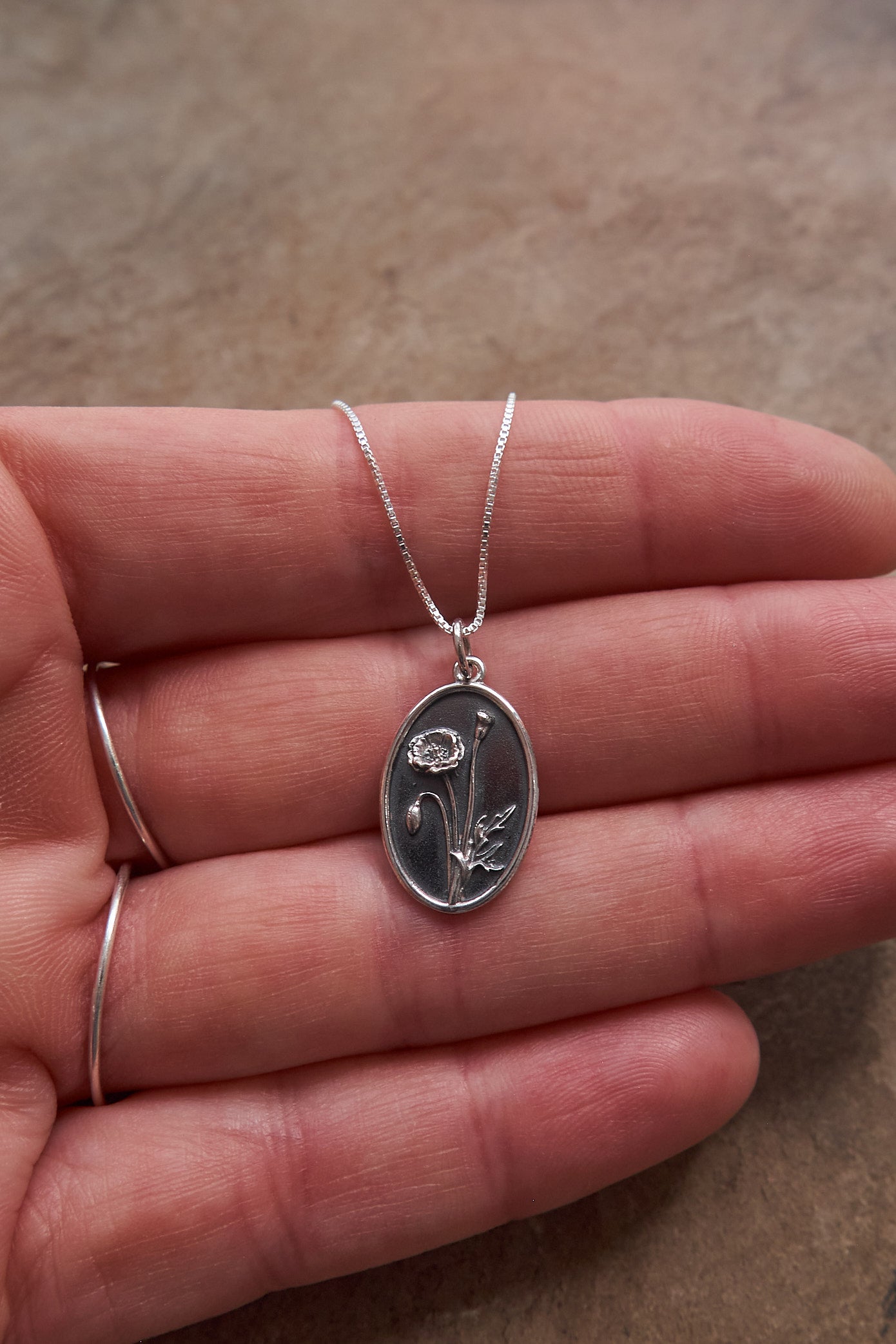 Silver poppy pendant necklace held in a hand on a neutral slate background
