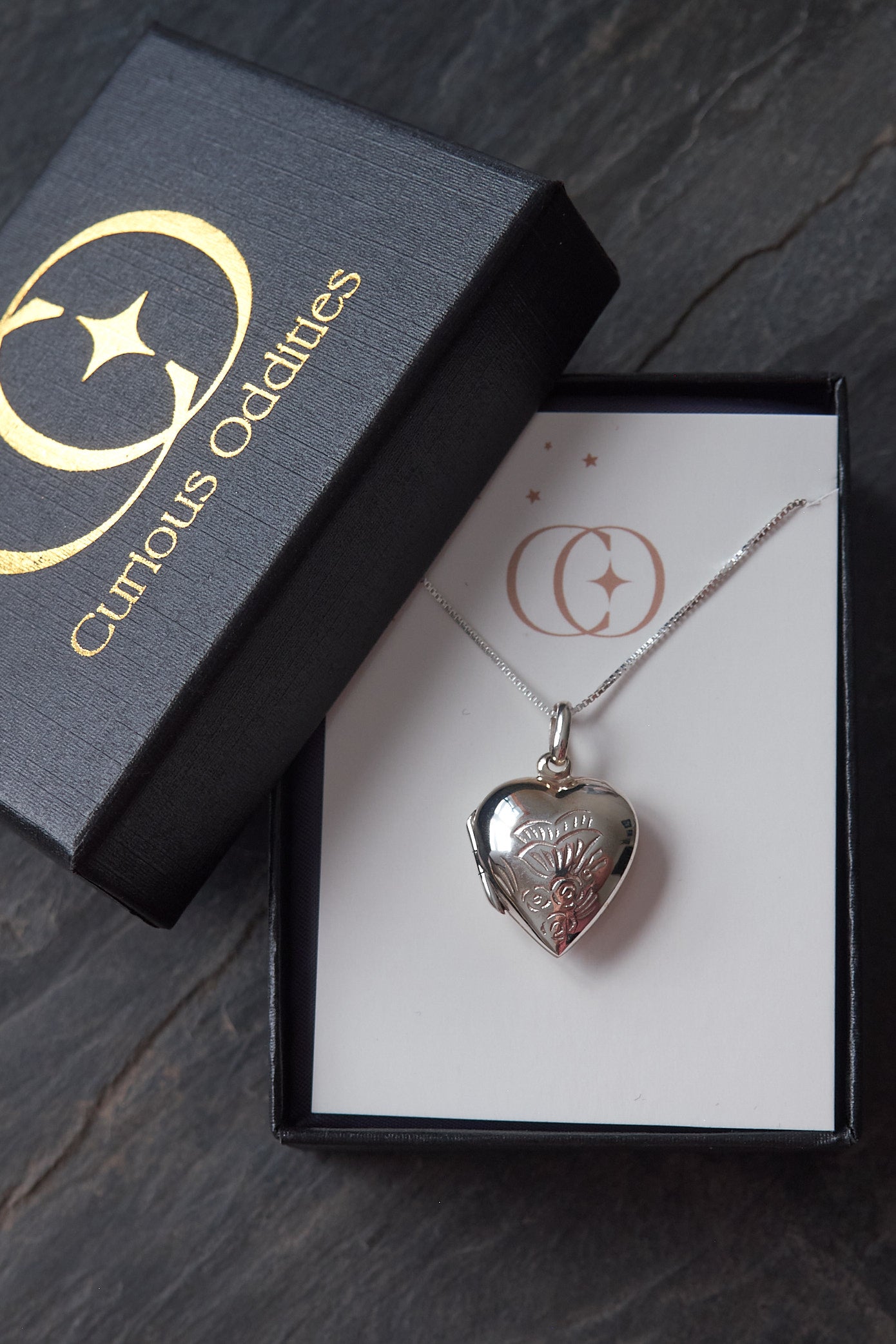 Silver heart-shaped locket in a box with 'Curious Oddities' branding on a slate surface.
