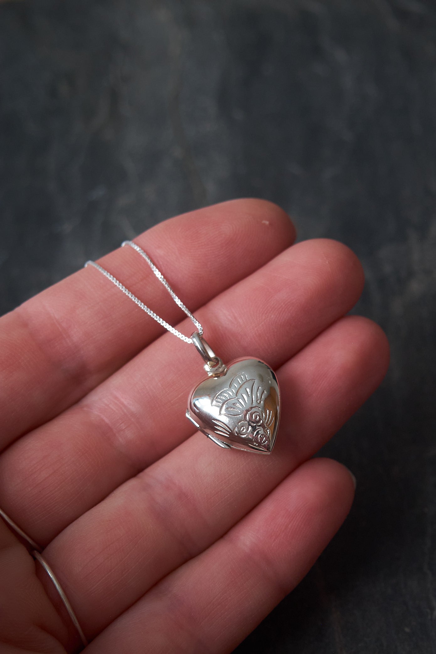 Silver heart-shaped locket on a chain held between fingers against a dark background