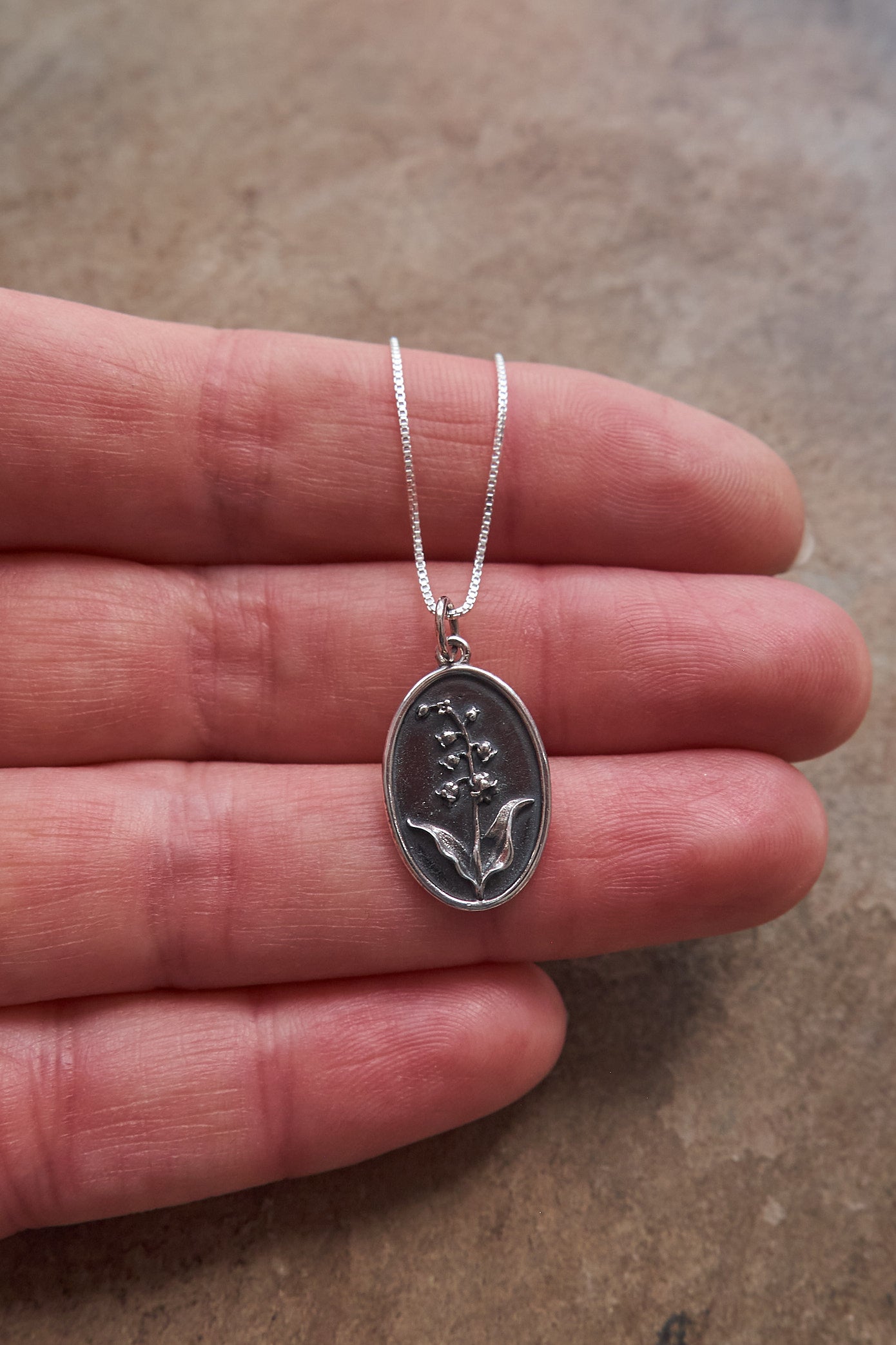 Silver necklace with a lily of the valley floral pendant held between fingers on a neutral background
