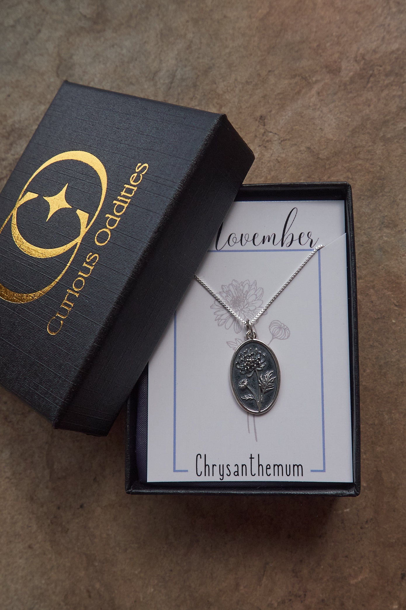 Silver necklace with chrysanthemum design in a 'Curious Oddities' box on a brown surface