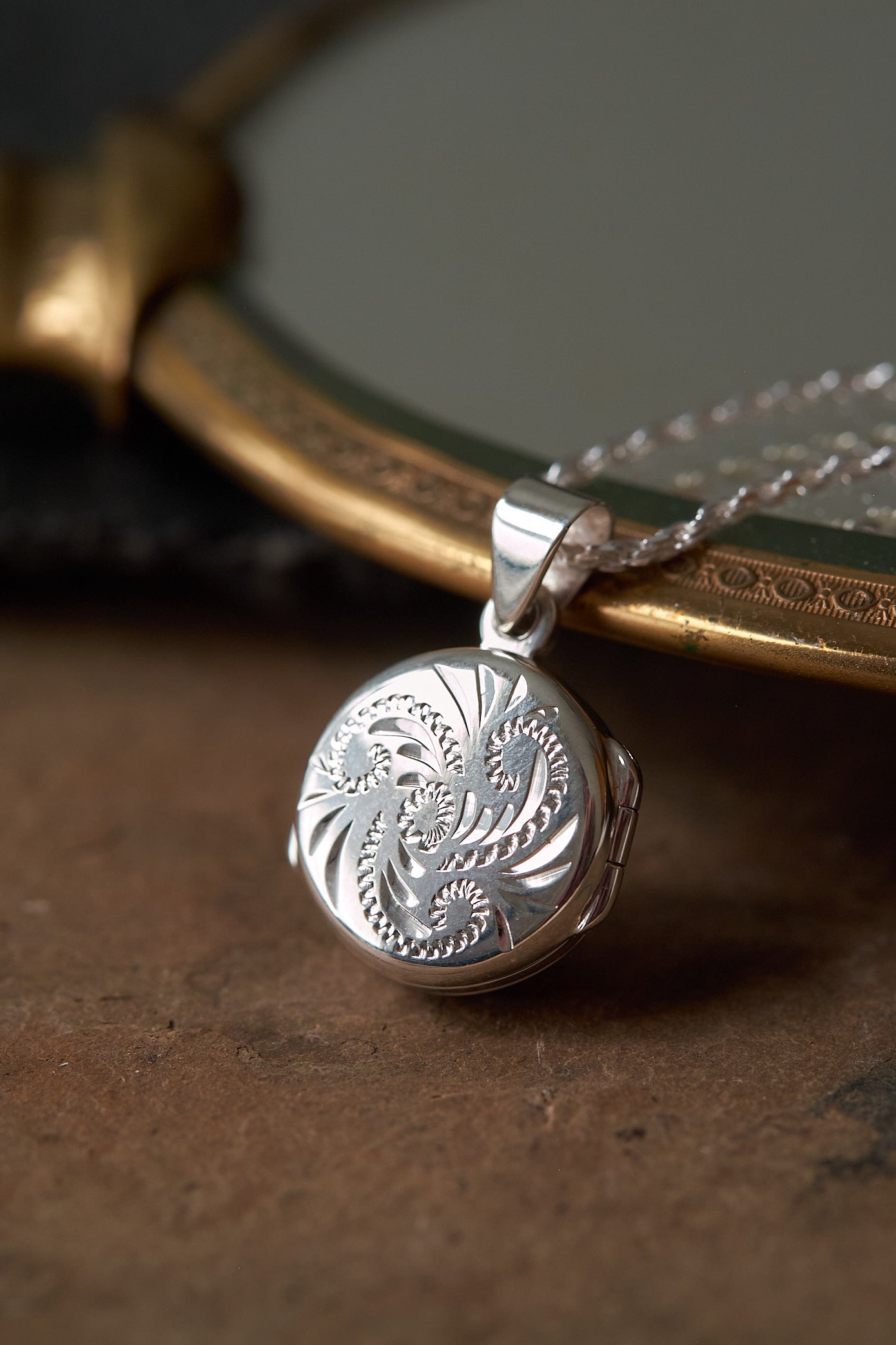 Silver locket with intricate swirl design on a chain, placed on a textured surface.